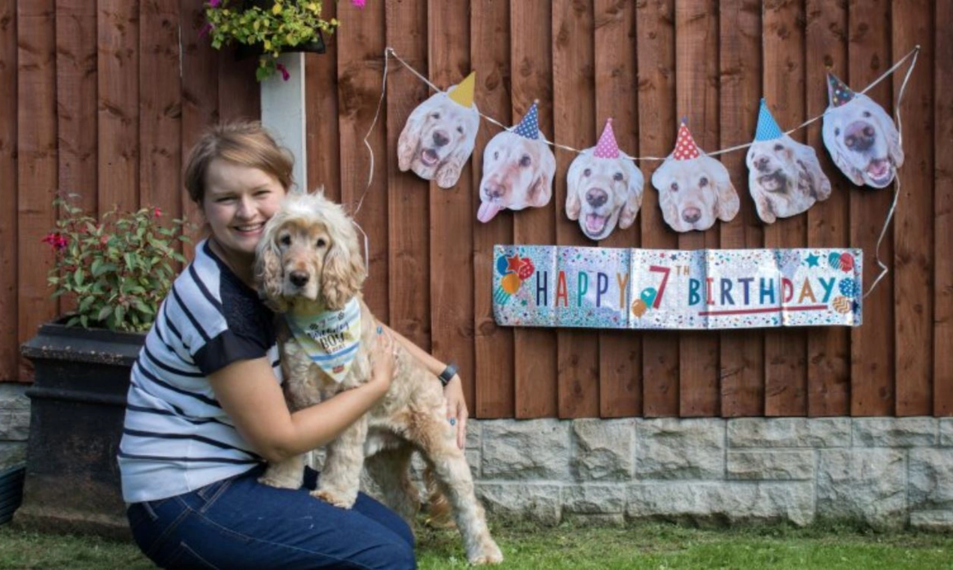A woman sat outside holding a dog with a 7th birthday banner in the background on a wooden fence