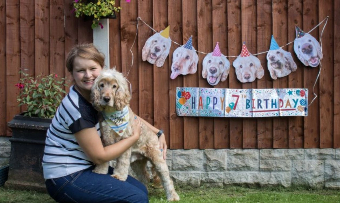 A woman sat outside holding a dog with a 7th birthday banner in the background on a wooden fence