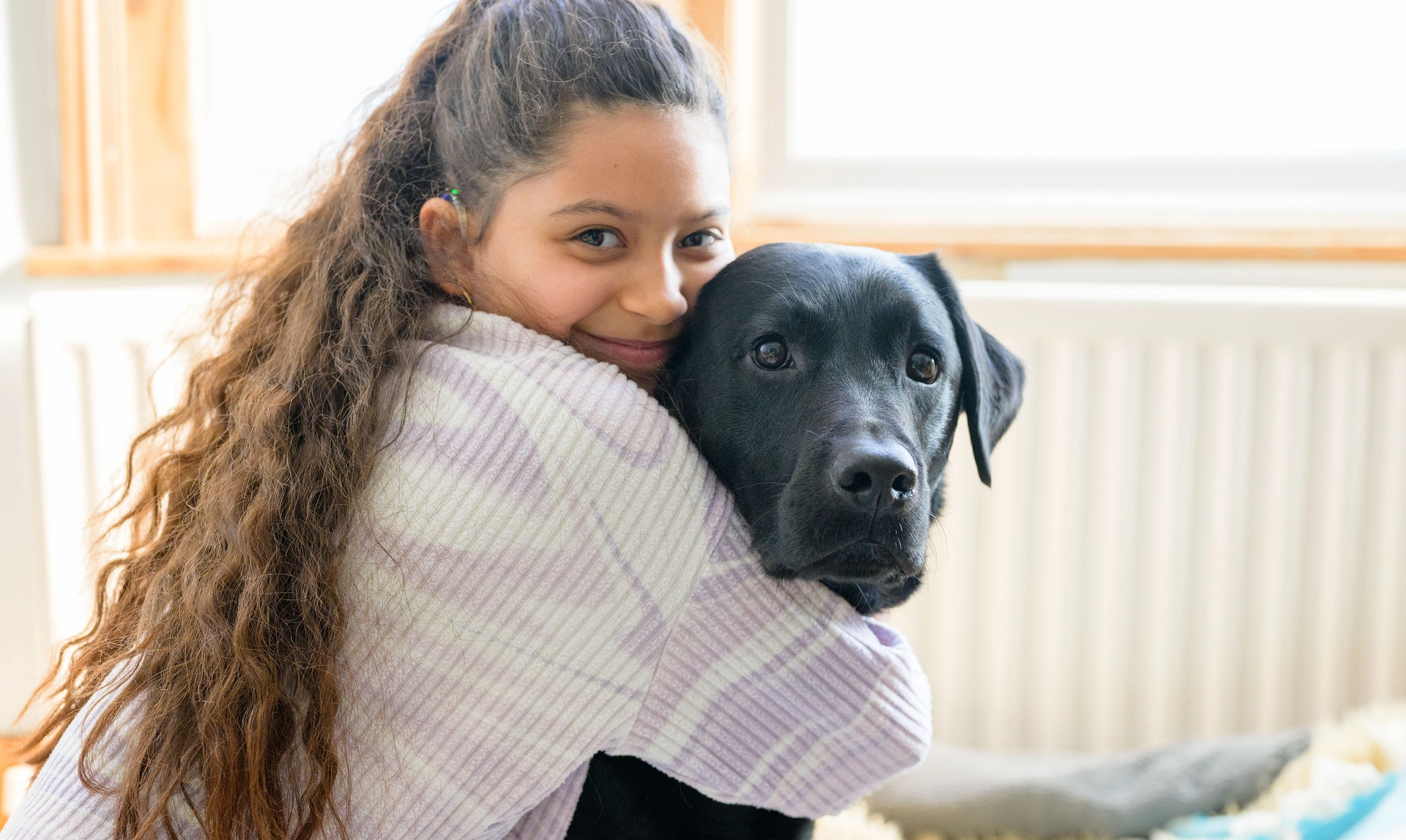 A young girl is hugging a black labrador and smiling while being sat in a house with a window and radiator in the background