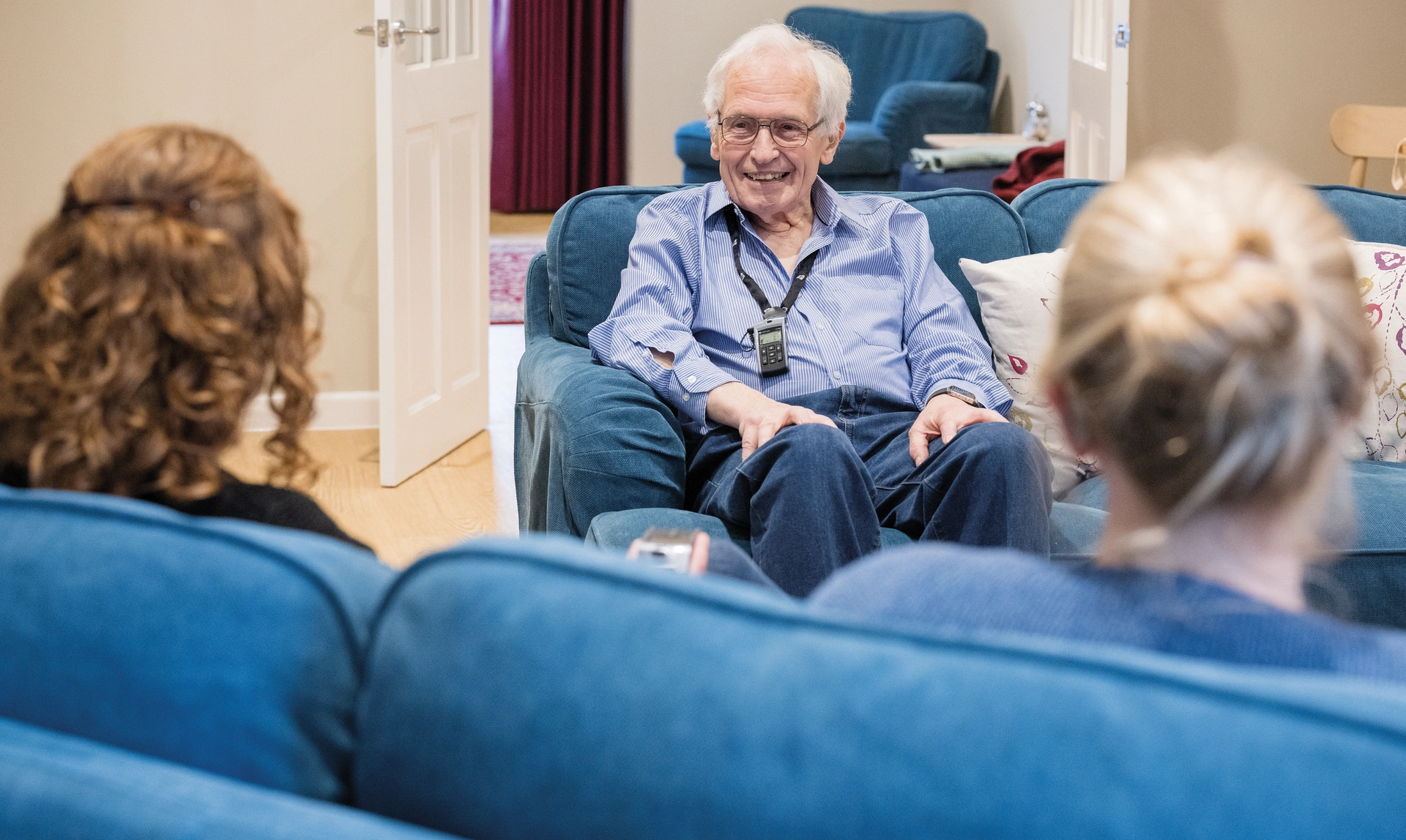 An older man with a hearing device around his neck sat opposite to two woman, they look like they are all talking