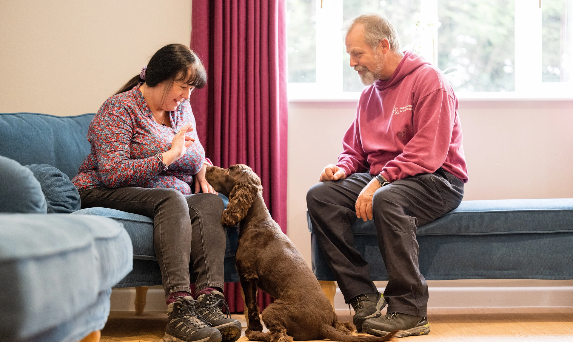 A woman is sat in a lounge with a man dressed in hearing dogs staff uniform meeting a brown spaniel