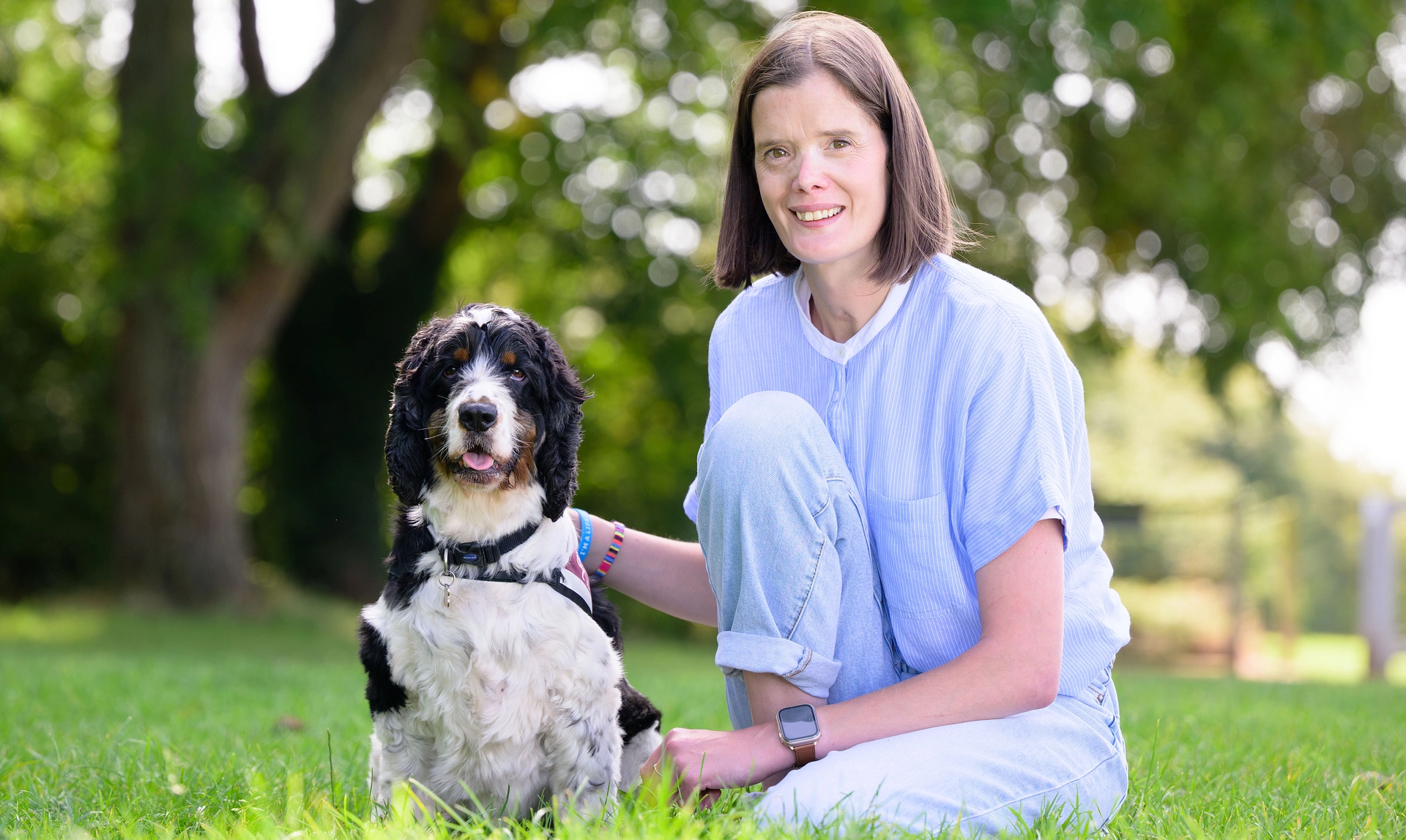 A woman sat on the grass smiling next to a black and white Cocker Spaniel, they are both looking directly at the camera, and sitting in front of large green trees