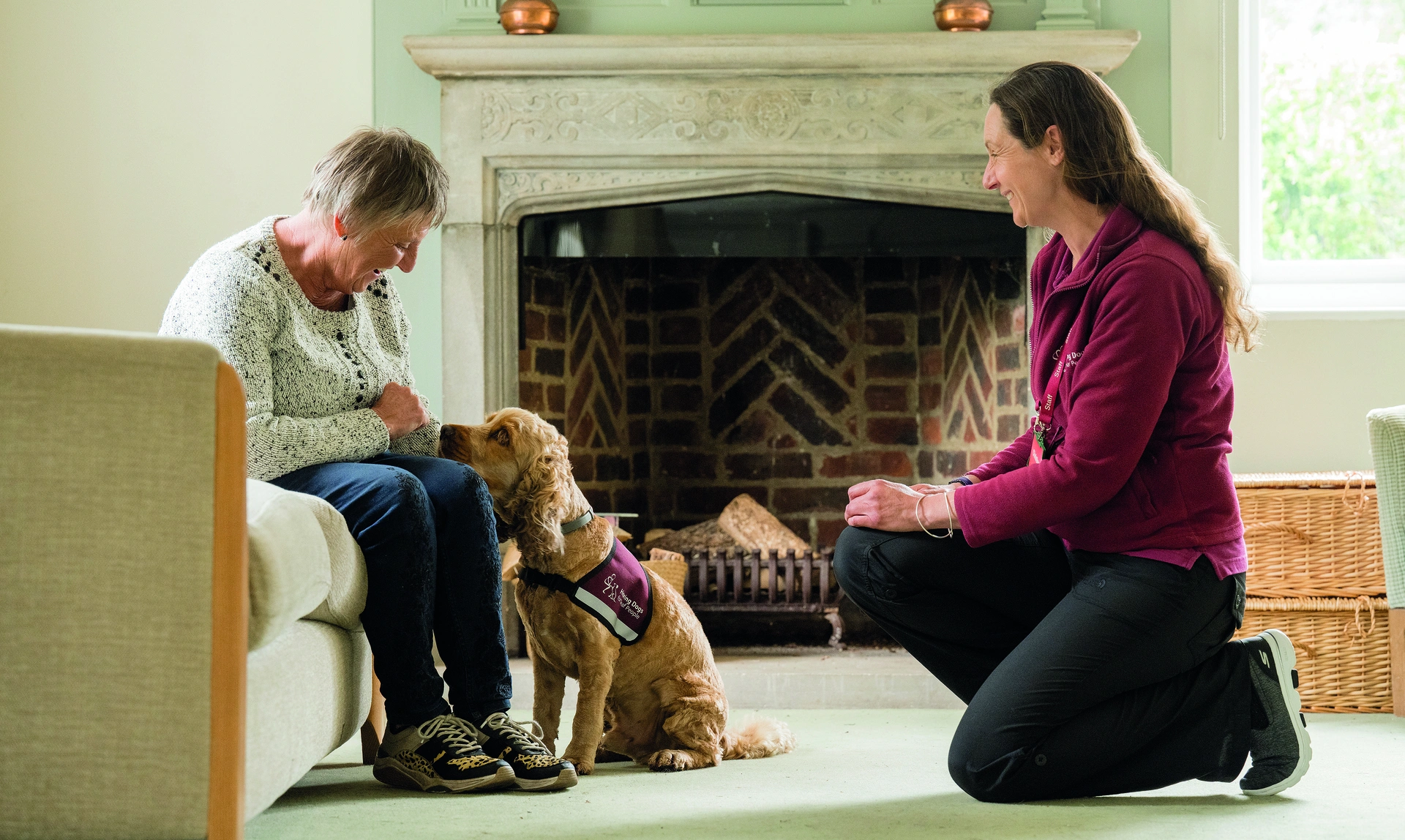 An older woman sat on a sofa looking happily at a Spaniel hearing dog which is looking up at her, there is a hearing dogs trainer crouched in front of them