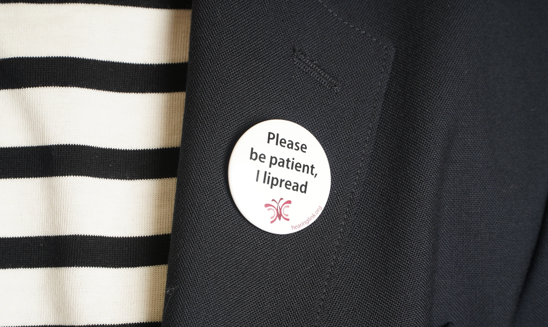Close up of a badge on a jacket lapel that says ‘Please be patient, I lipread’
