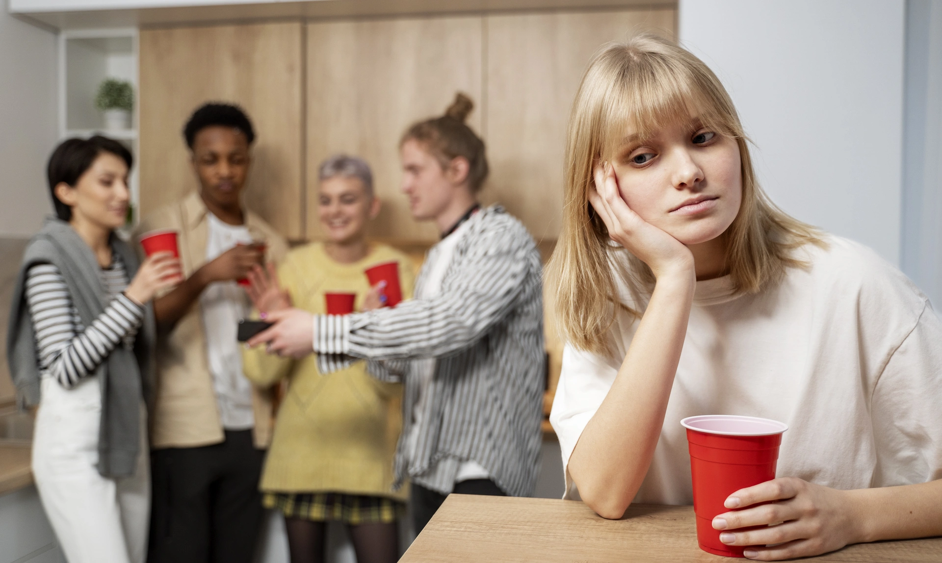 Woman in foreground sat at a table alone looking unhappy while four other people are talking happily in the background
