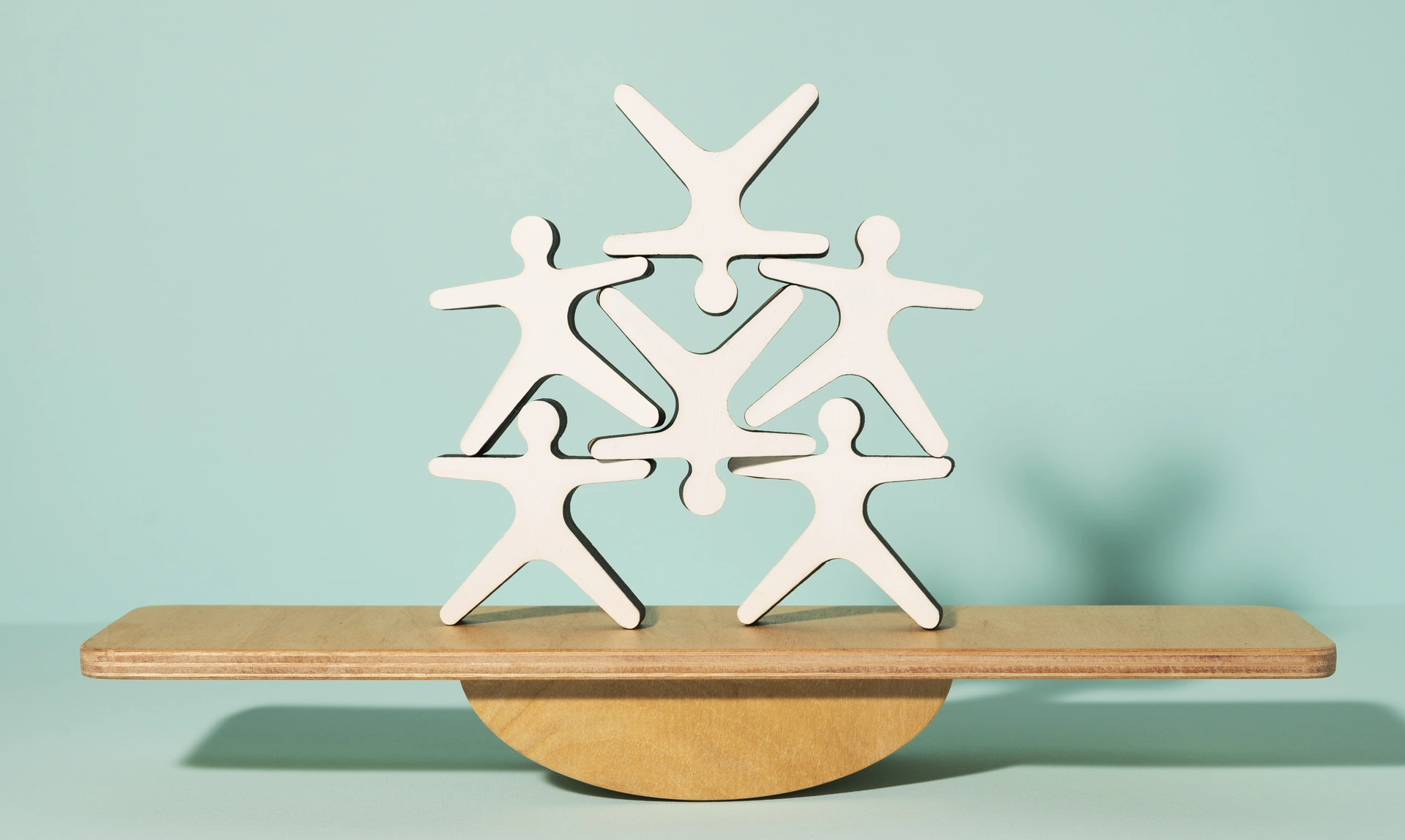 Paper cutouts of people stacked on top of each other on a small wooden seesaw