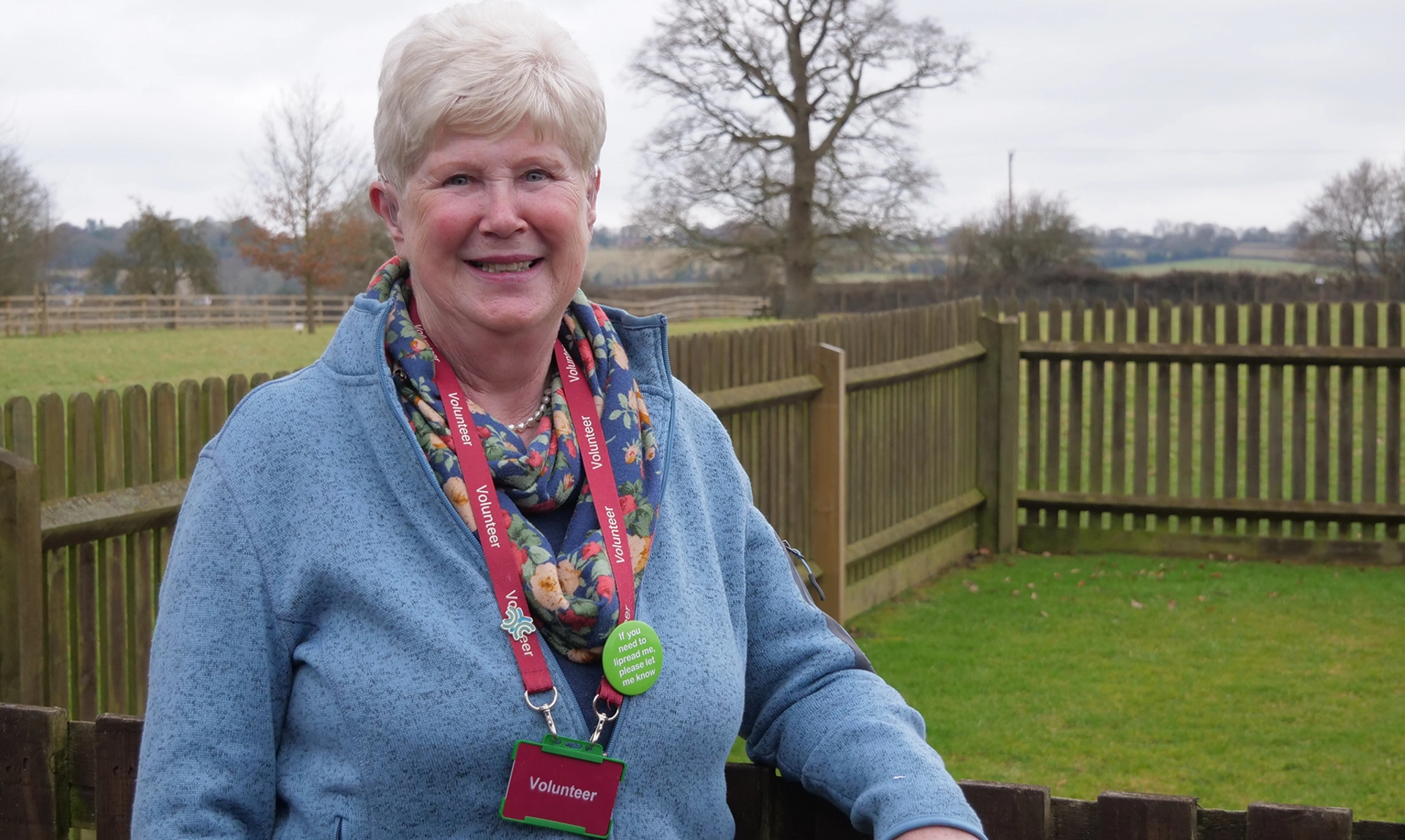 Mid shot of a happy woman leaving on a fence outside looking directly at the camera and wearing a lanyard with a badge that says ‘Volunteer’
