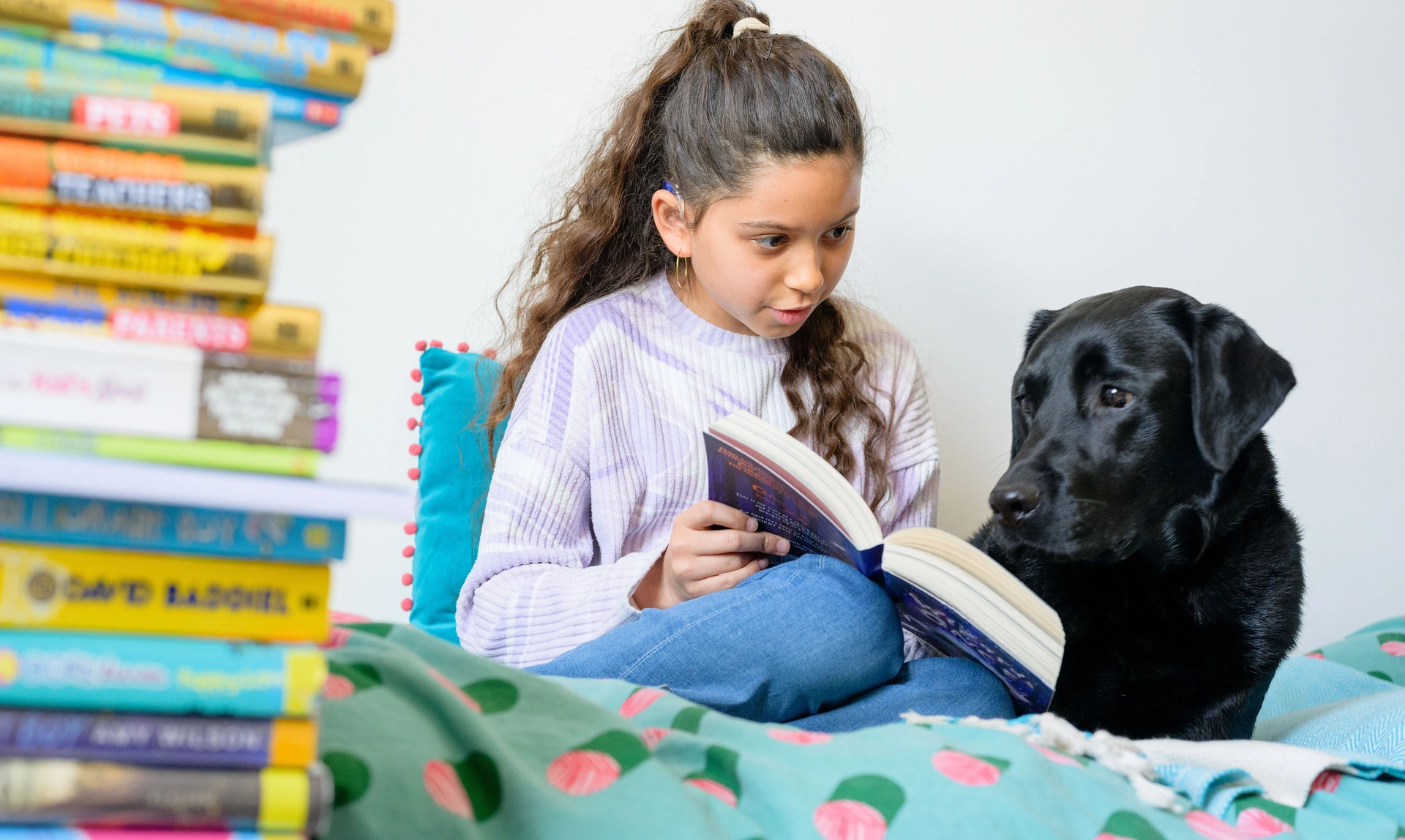 A young girl sat on her bed with a pile of books, happily reading to a labrador