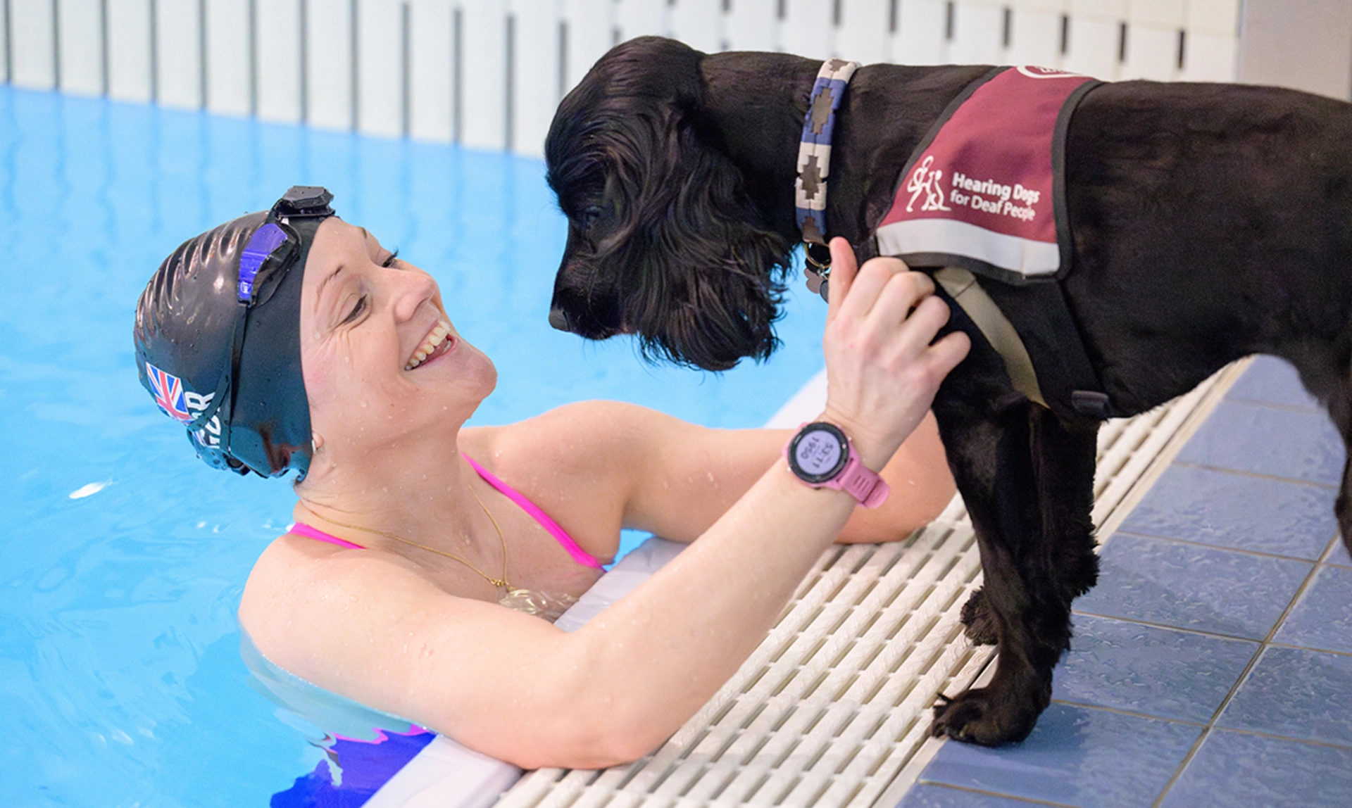 A woman in a swimming pool with a hearing dog in a jacket at the poolside looking at her