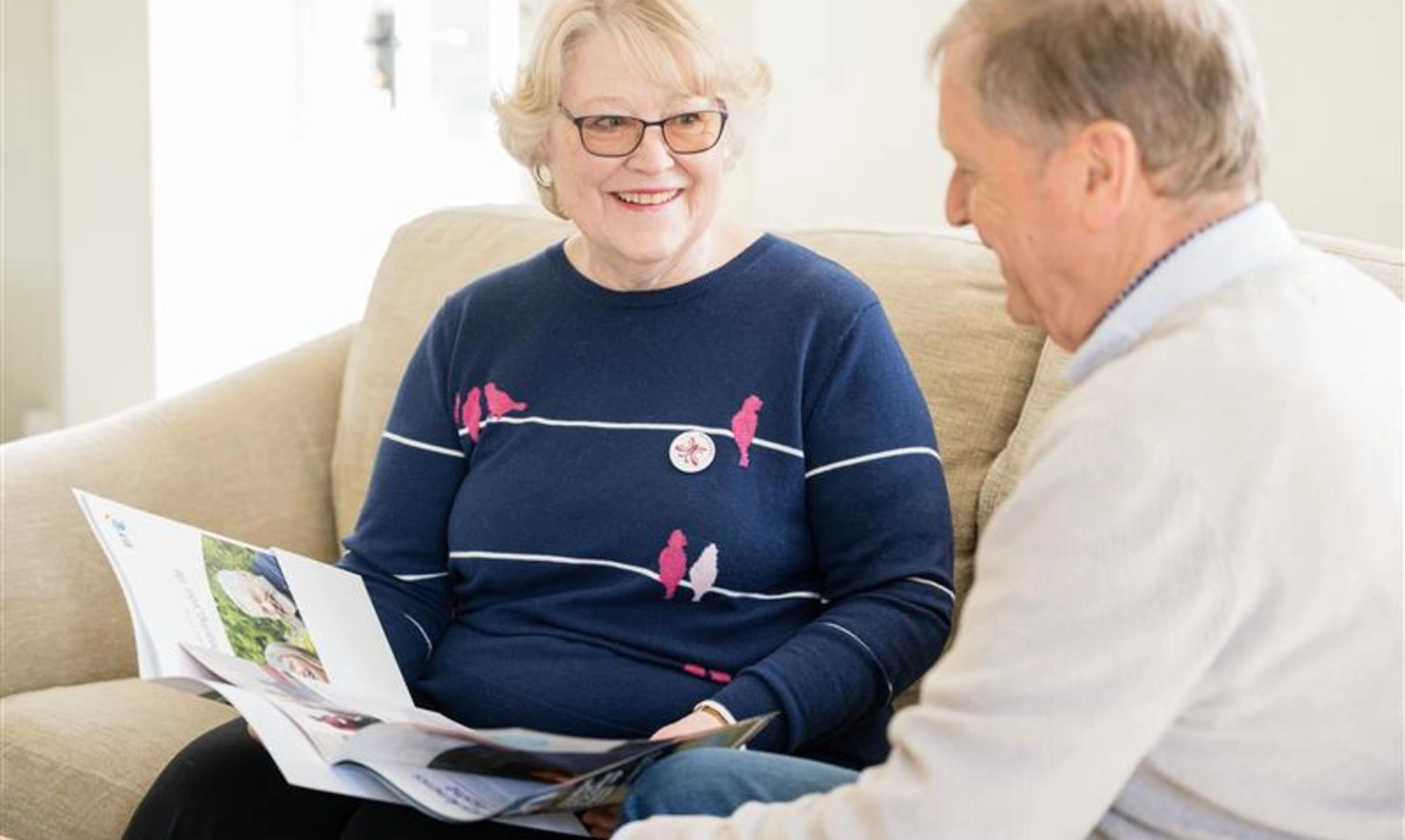A woman and man sitting on sofa. The woman is showing the man a book.