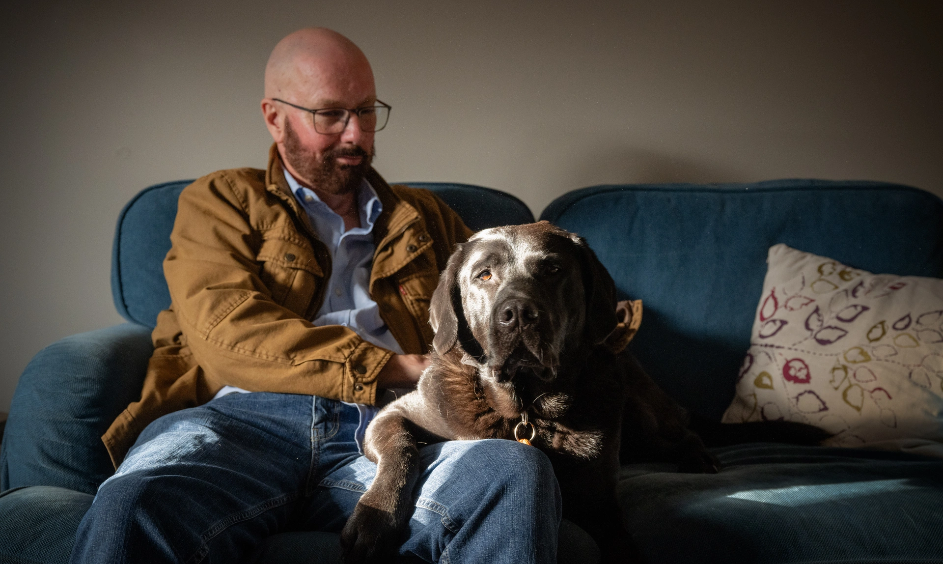 A bearded man, Trevor,  sitting on a sofa with a large brown dog on his lap.