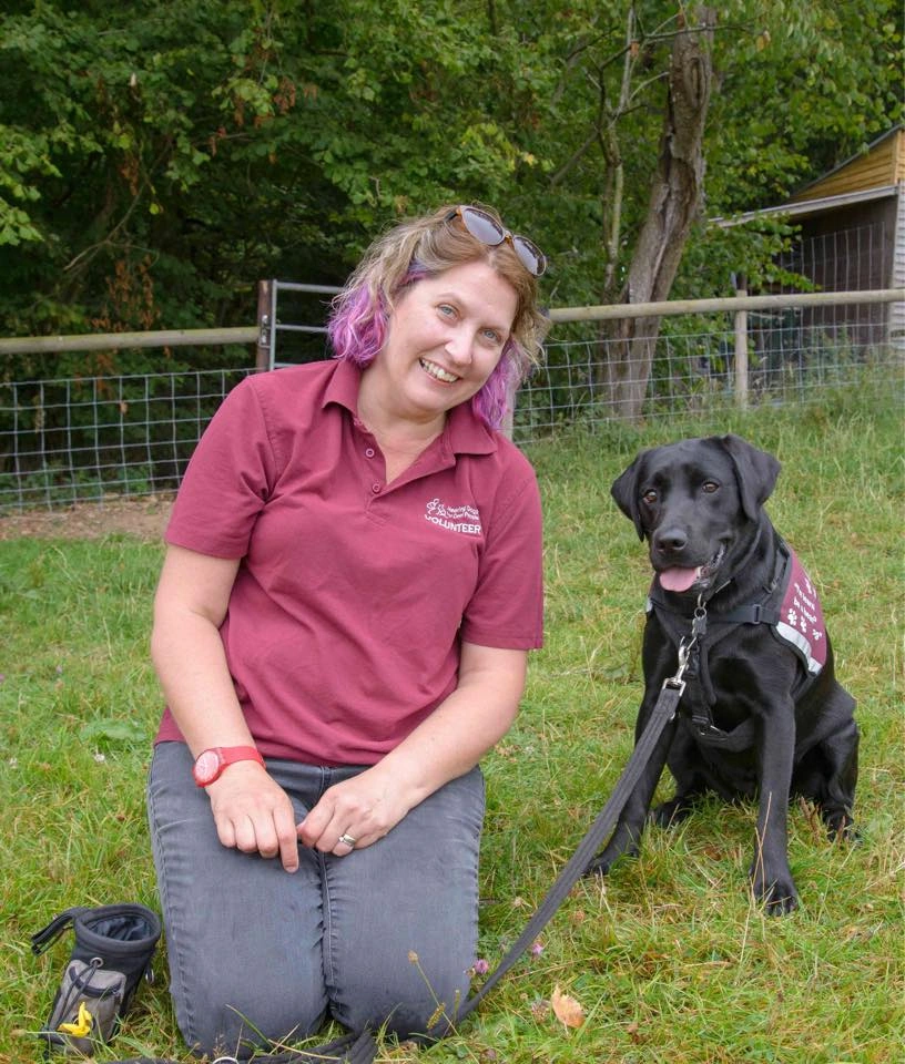 Woman sat on grass in burgundy hearing dogs tshirt next to black labrador in their hearing dogs jacket