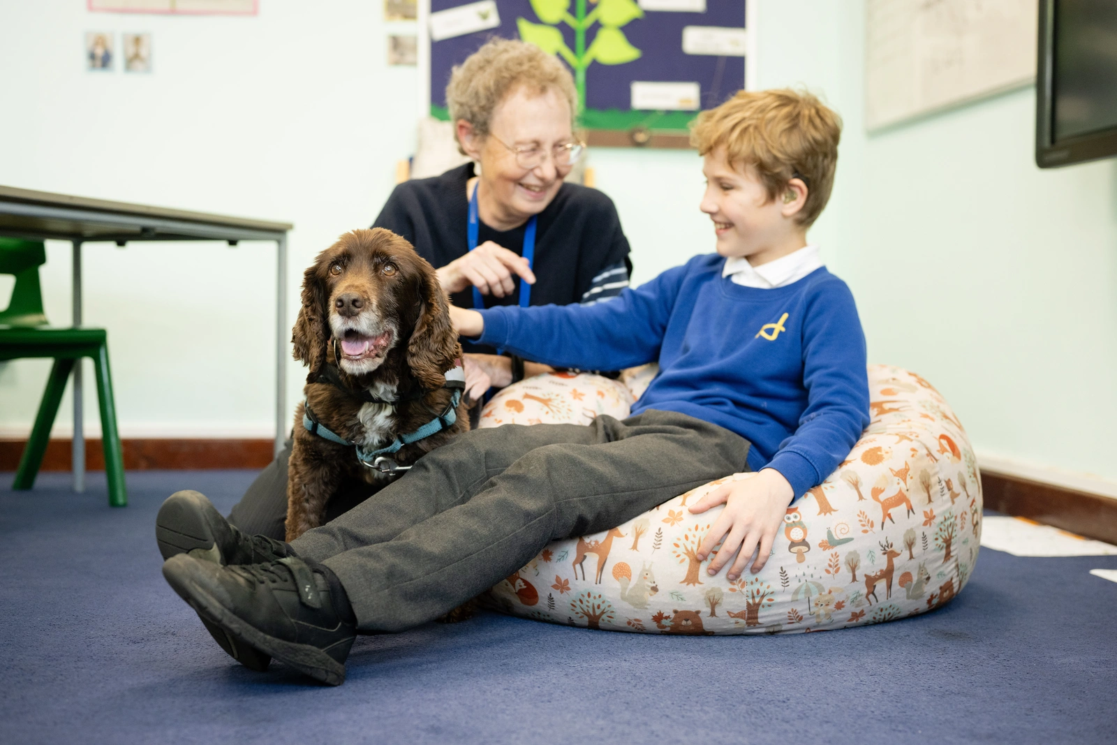 A young boy in blue uniform jumper is seated on a bean bag chair. He is stroking a brown down as a woman kneels smiling beside them
