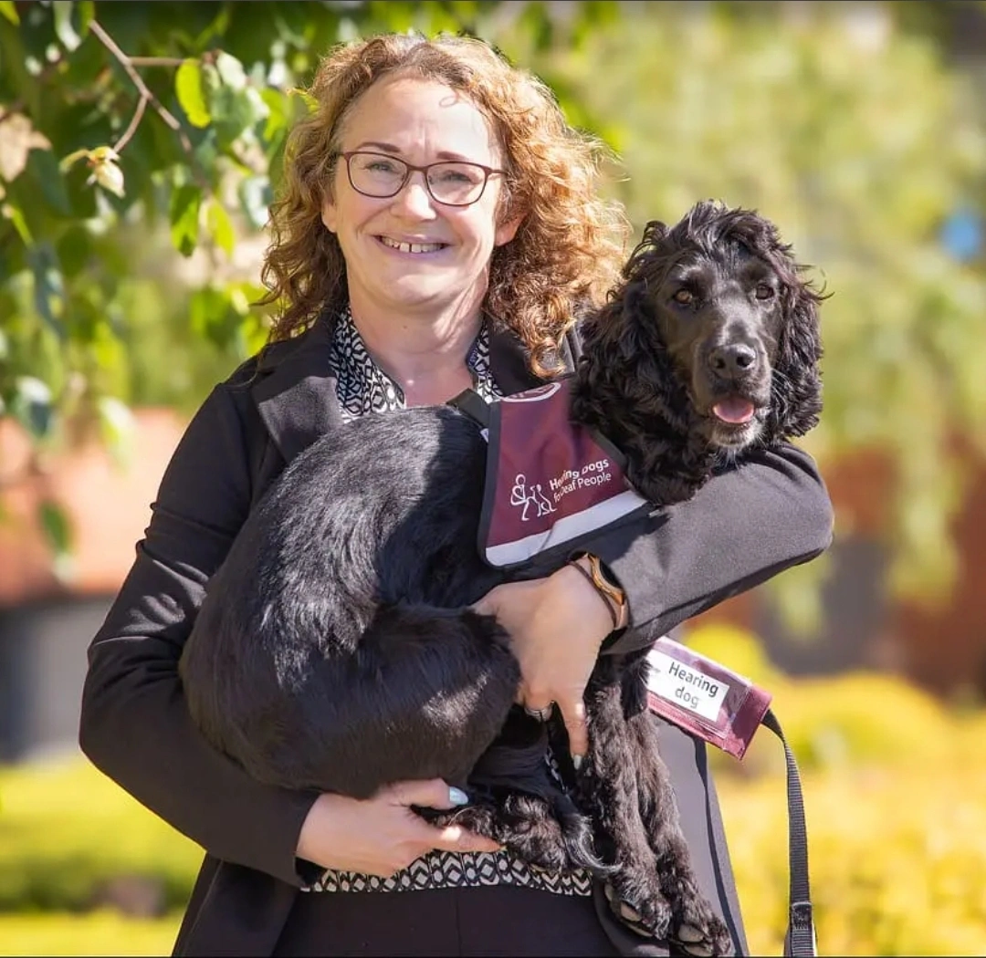 a woman smiling holding a black spaniel in a bugundy hearing dog jacket