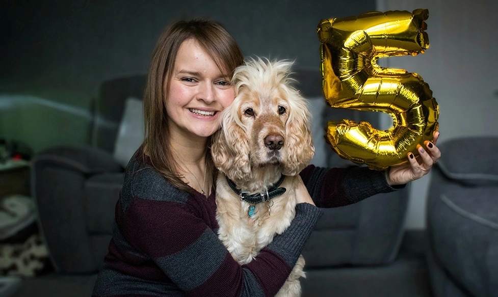 A smiling woman is hugging a light sandy dog, while holding a metallic balloon in the shape of a number five.