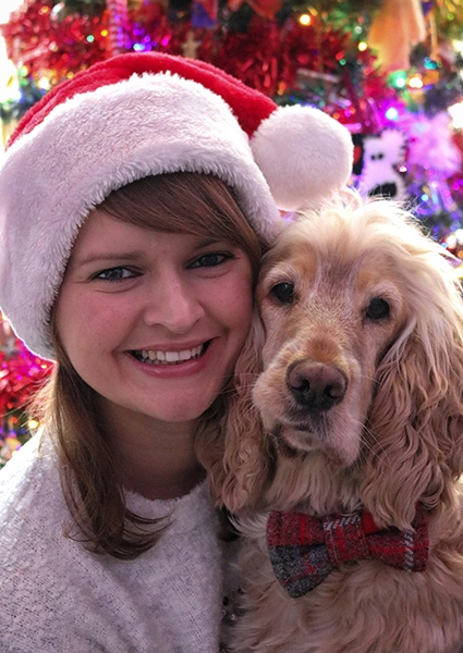 A woman wearing a Christmas santa hat, next to a light coloured dog that is wearing a bow tie. A Christmas tree can be partially seen in the background.