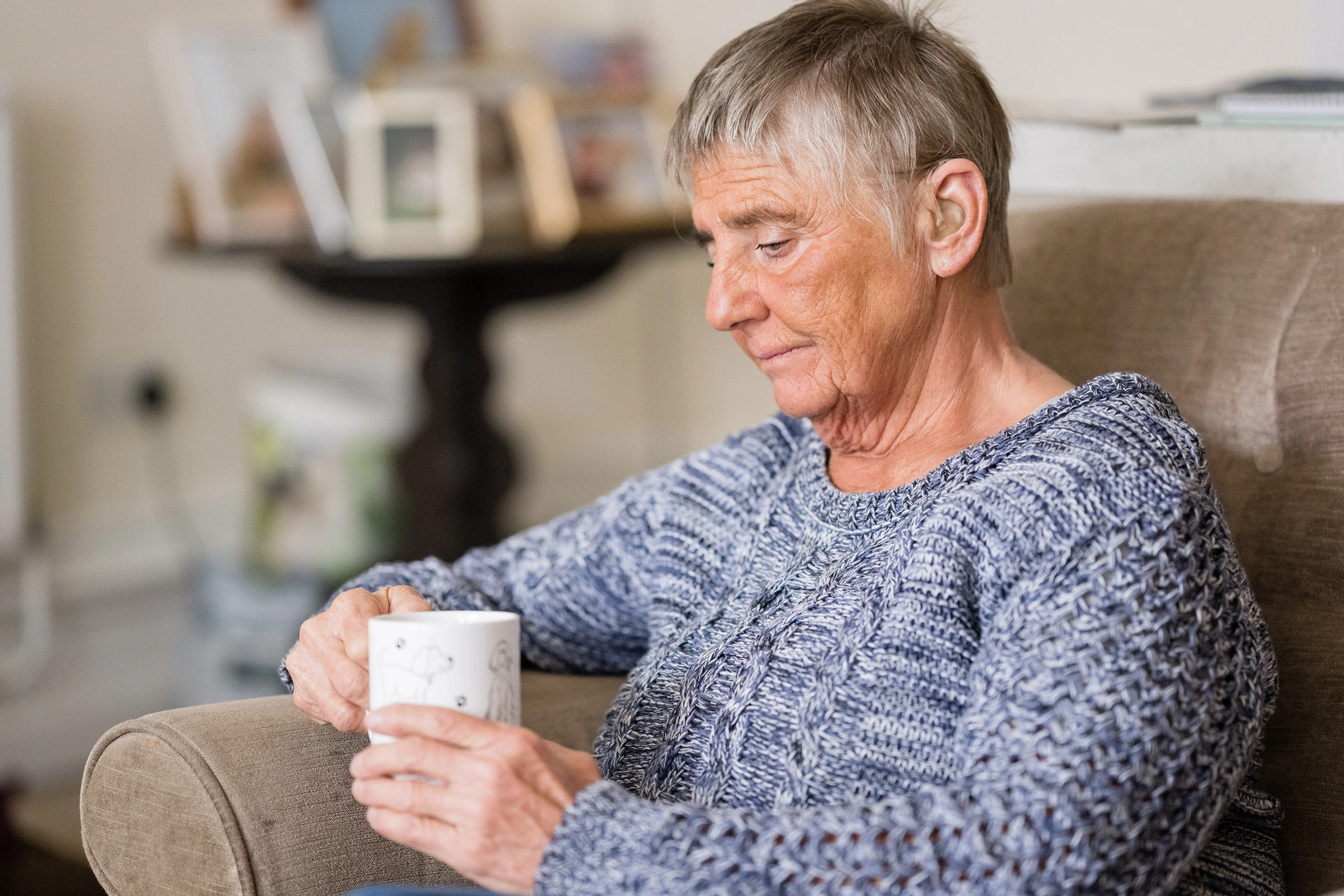 A woman with short greying hair sitting down holding a mug, She looks unhappy and saddened.
