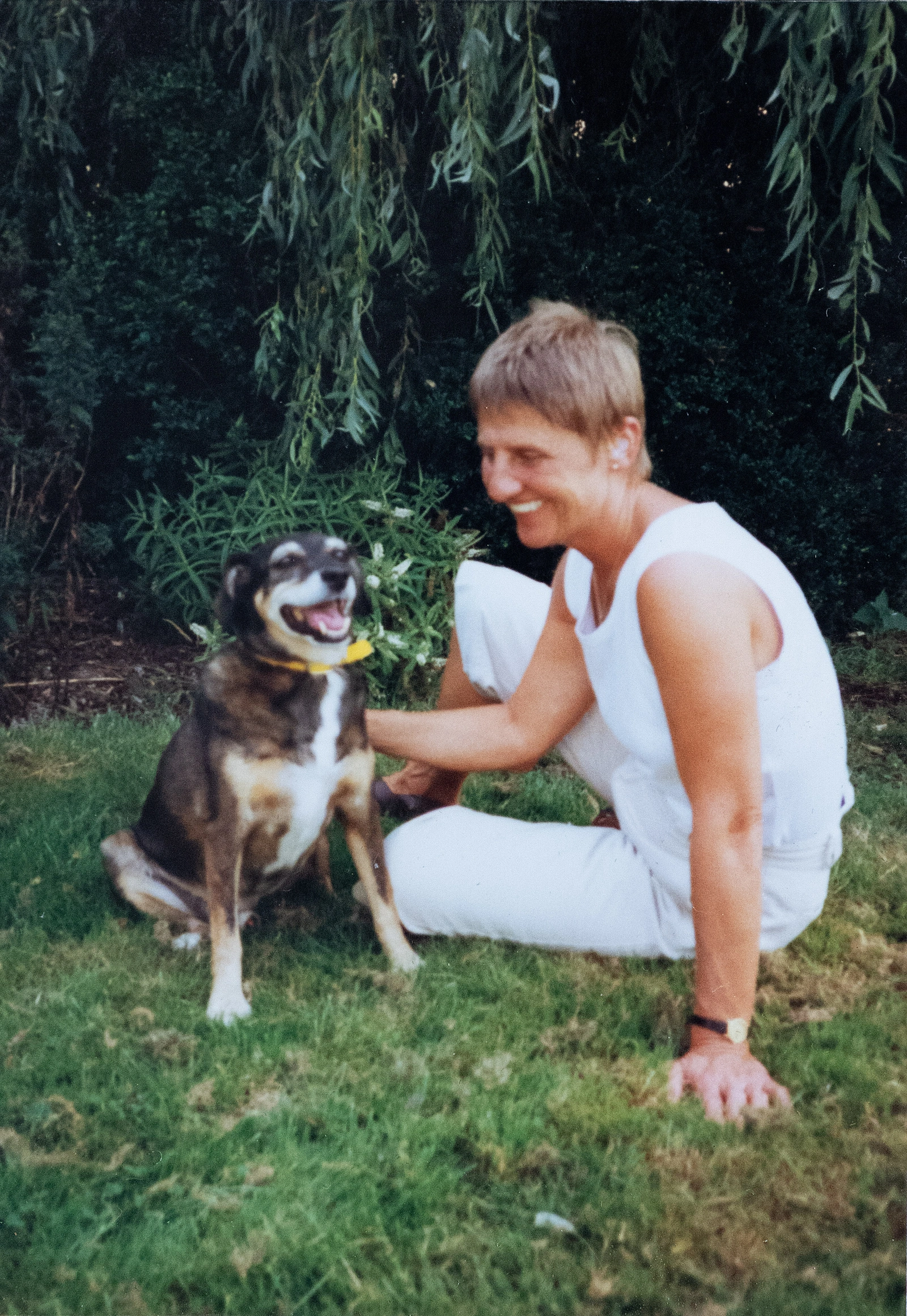 The woman from the previous pictures when younger, wearing white shorts and top. She is sitting on grass beside a brown and white dog, smiling broadly.
