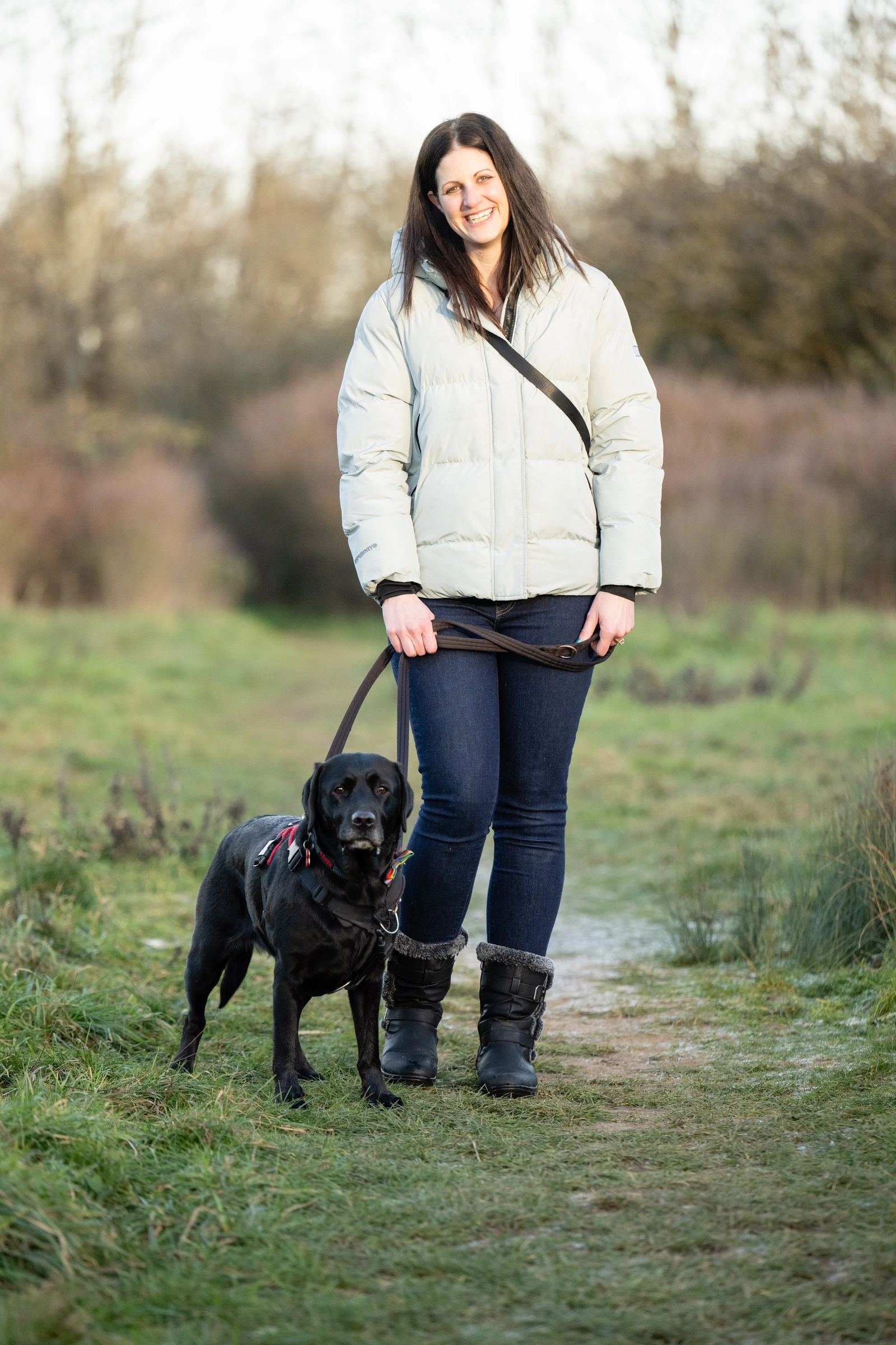 A woman wearing a white jacket and a black labrador standing in a field. The woman is smiling as the dog looks aheadnintently.