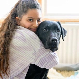A young girl is hugging a black labrador and smiling while being sat in a house with a window and radiator in the background