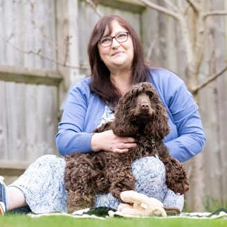 A bespectacled woman wearing a blue cardigan over a long dress is sitting on grass holding a brown curly haired dog in her arms.