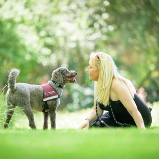 A blonde woman and a dog are in a grassy, tree filled location. She is sitting on the grass looking at the dog who is looking back at her, tail wagging.