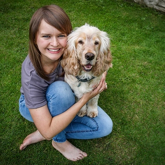 A young woman and light coloured dog are sitting on grass. The woman is smiling and hugging the dog.