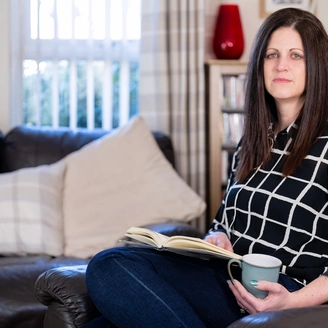 A woman with dark shoulder length hair is sitting on a dark sofa in a living room holding a book in her lap. She has a serious look on her face.