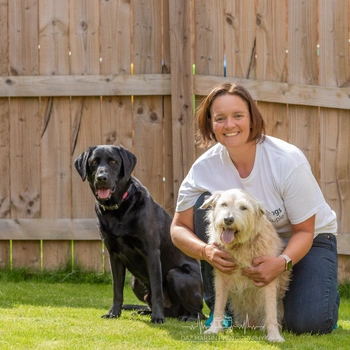 A young woman crouched next to a fluffy light-coloured dog and a black Labrador smiling at the camera in the sunshine with a garden fence behind them