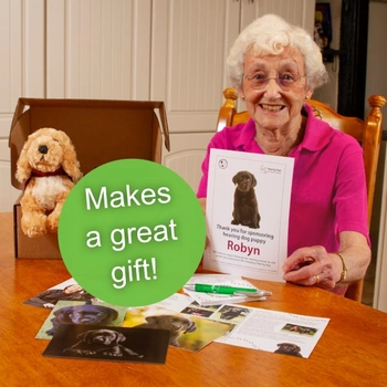 Older adult woman in a bright pink top sat at a wooden table holding up a certificate with a black Labrador puppy on it and the name Robyn, the rest of the welcome pack is also on the table