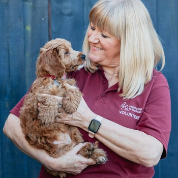 A blonde woman wearing a hearing dogs volunteer tshirt is standing smiling while holding a small puppy