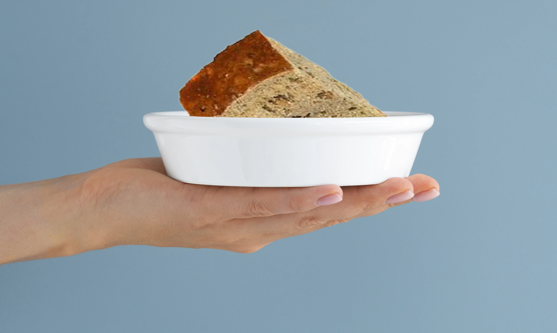  A hand holding up a white dog owl containing square serving of grey brown cake which is darker on the top, the background is teal colour