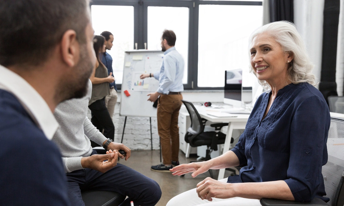 Three people sat talking in an open plan office with other people in the background