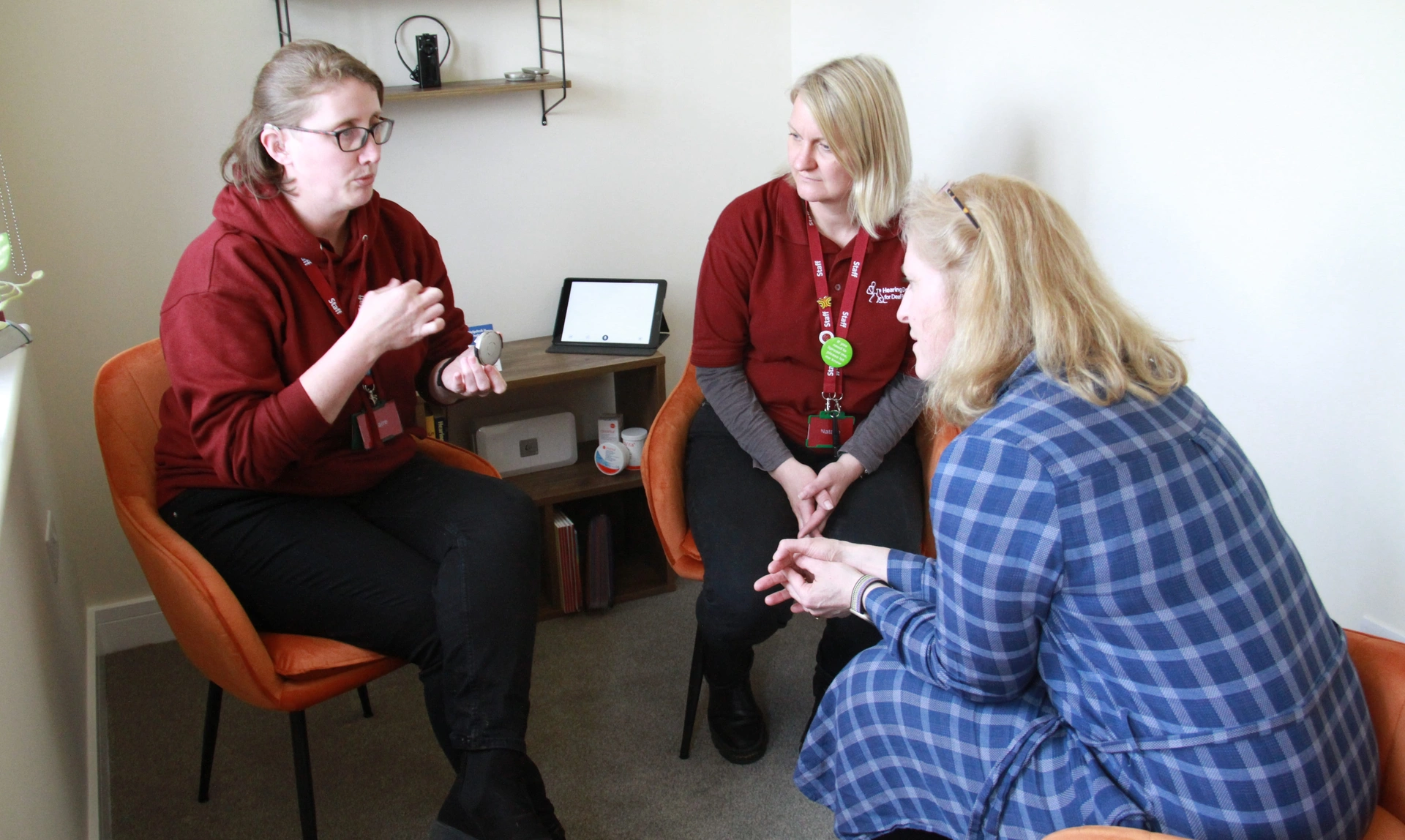 Two women dressed in hearing dogs uniform sat in a small room with another woman holding up a hearing device
