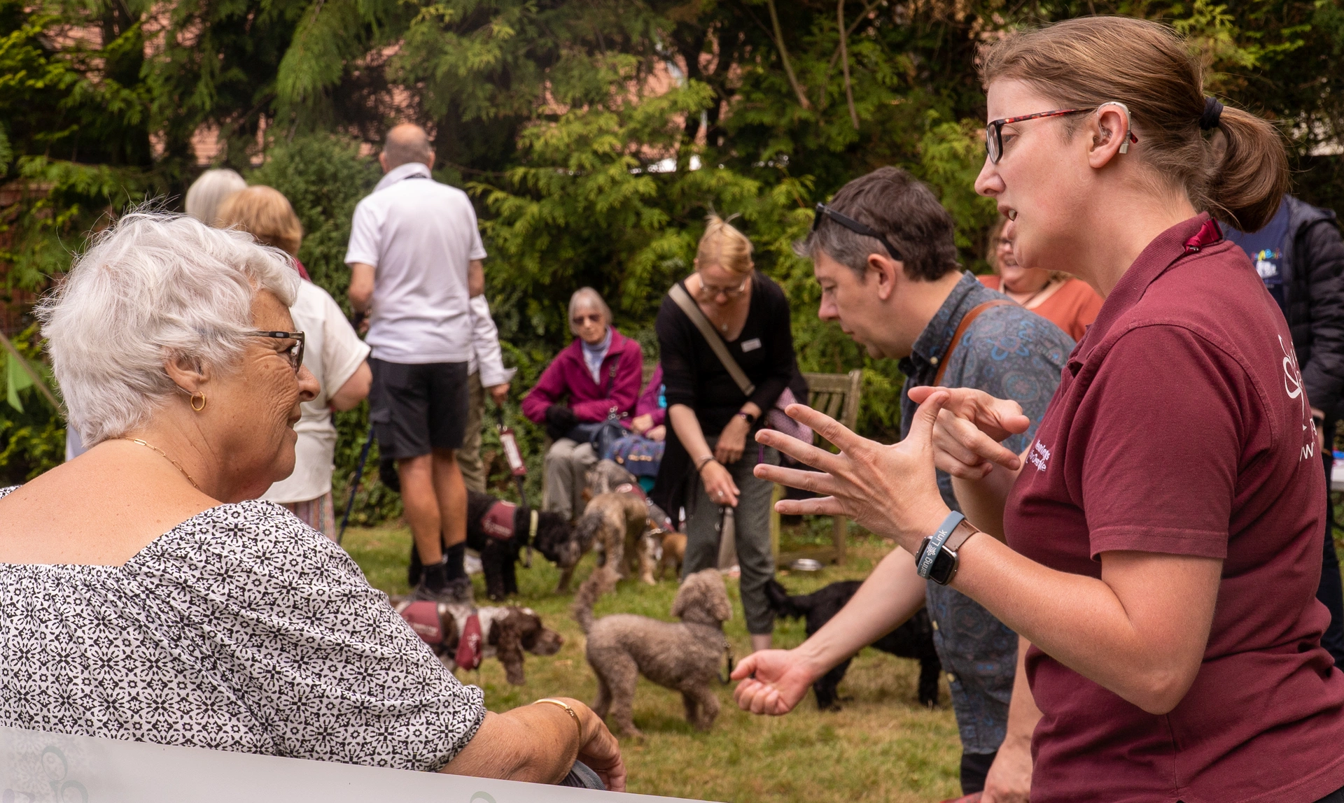 One woman using BSL in a hearing dogs uniform talking to another woman, outdoors with other people in the background