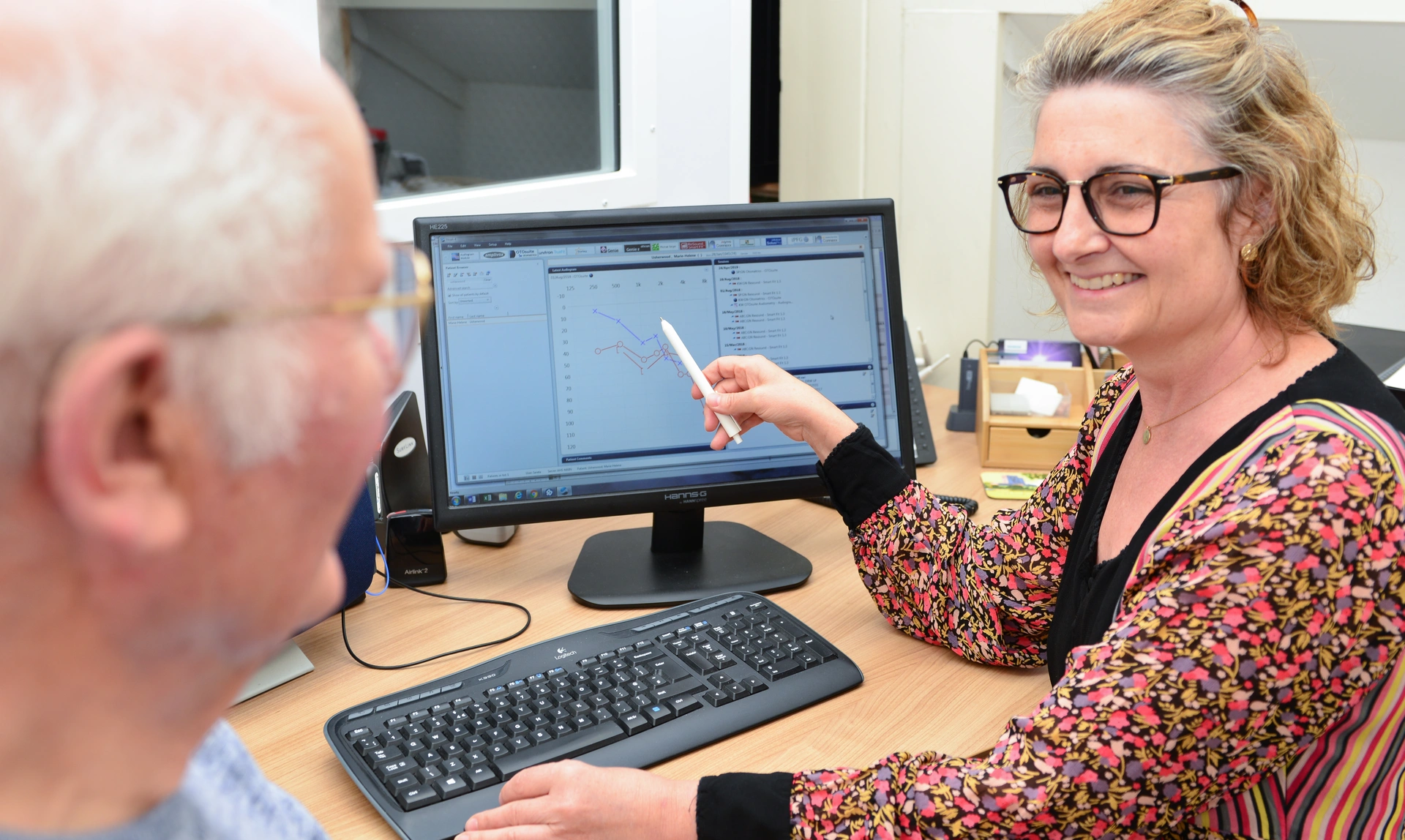 A female audiologist pointing to hearing test readings on a computer screen and smiling as an older man sat with her