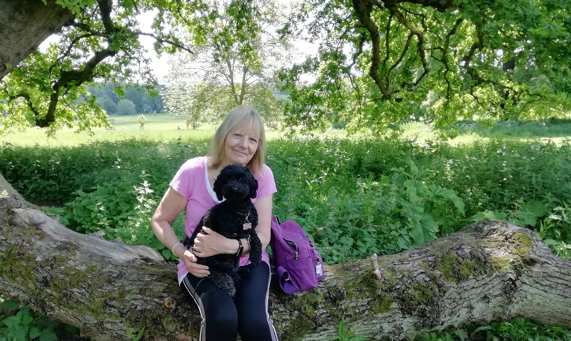 A woman in woods sat on a log with her black poodle on her lap