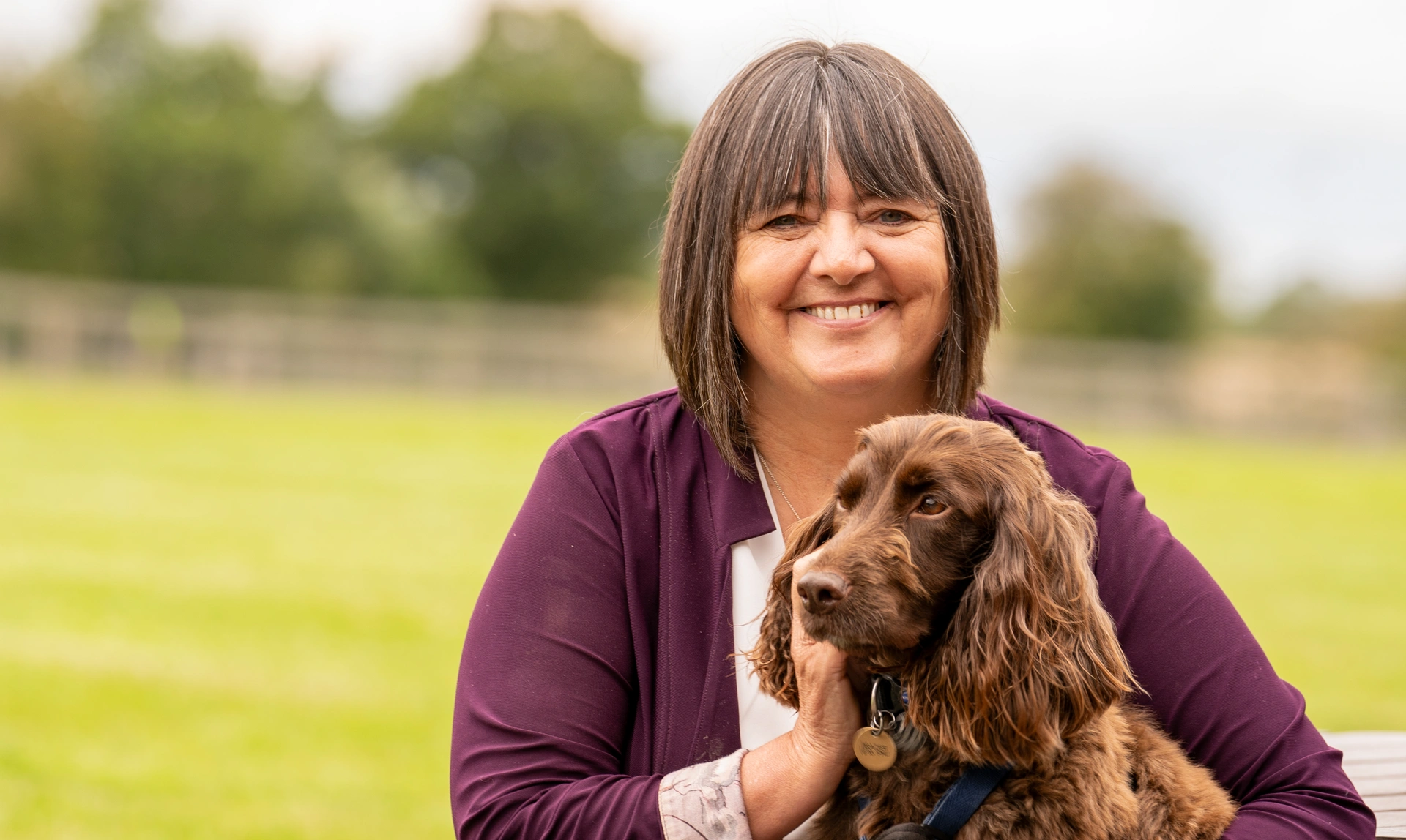 A woman sat outside smiling at the camera with a brown Spaniel sat on her lap