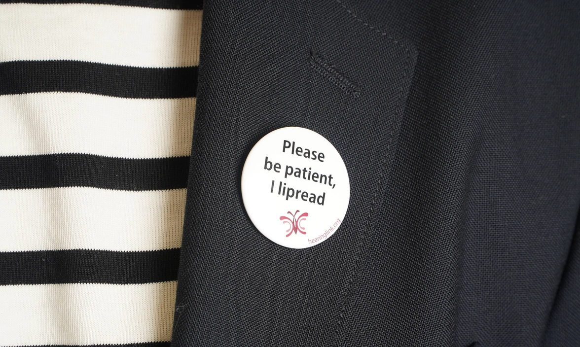 Close up of a badge on a jacket lapel that says ‘Please be patient, I lipread’