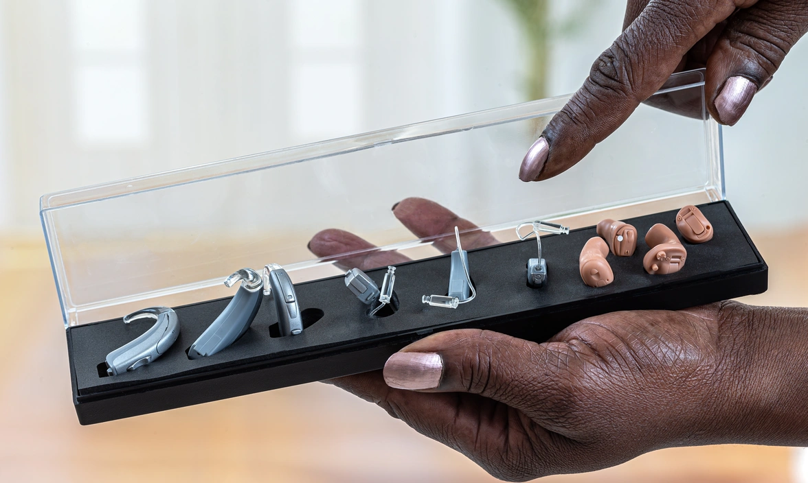 Close up of one hand holding a display case of four different types of hearing aid while the other hand points at them