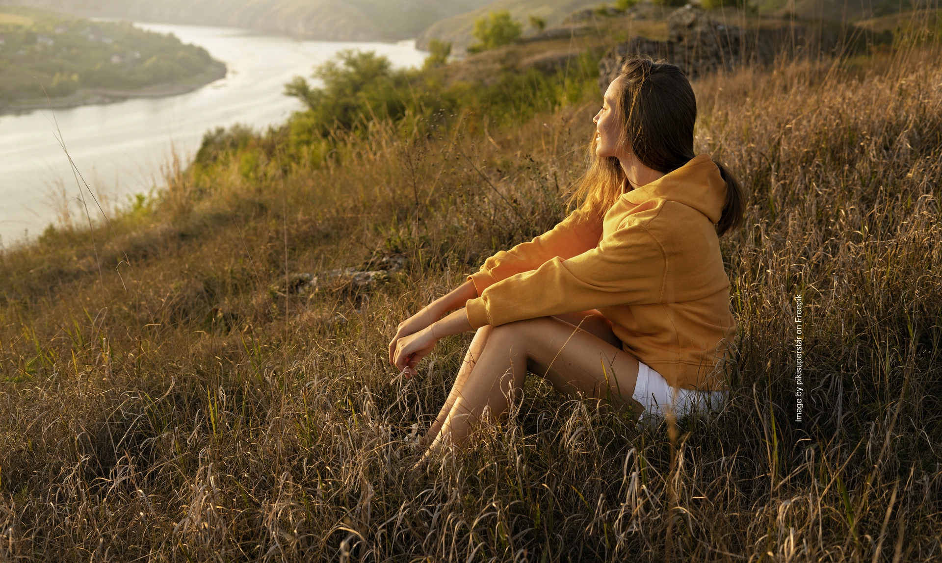 A woman sat on a hill as the sunsets looking out at some countryside