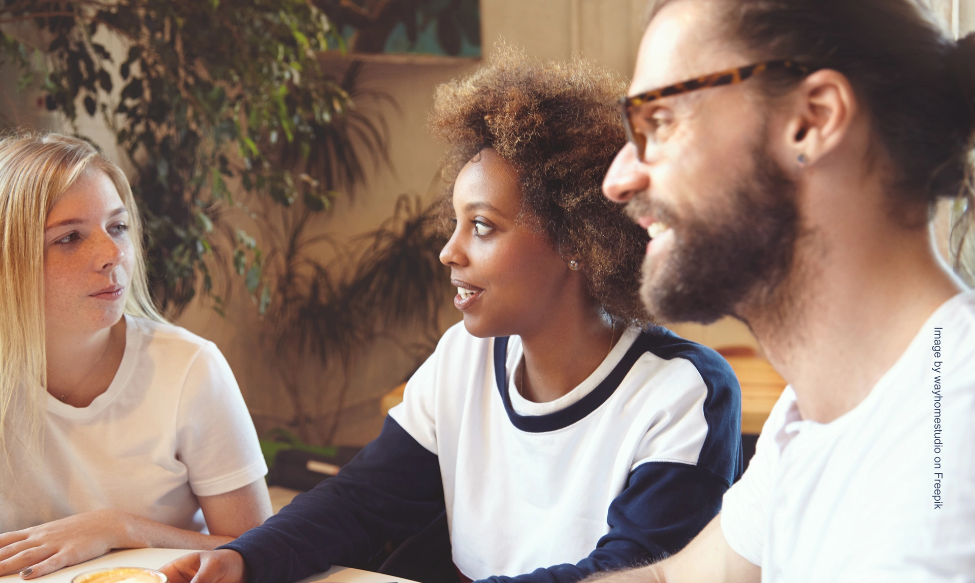 Mid shot of three people sat at a table communicating, two of them are listening and smiling as they look at the other person