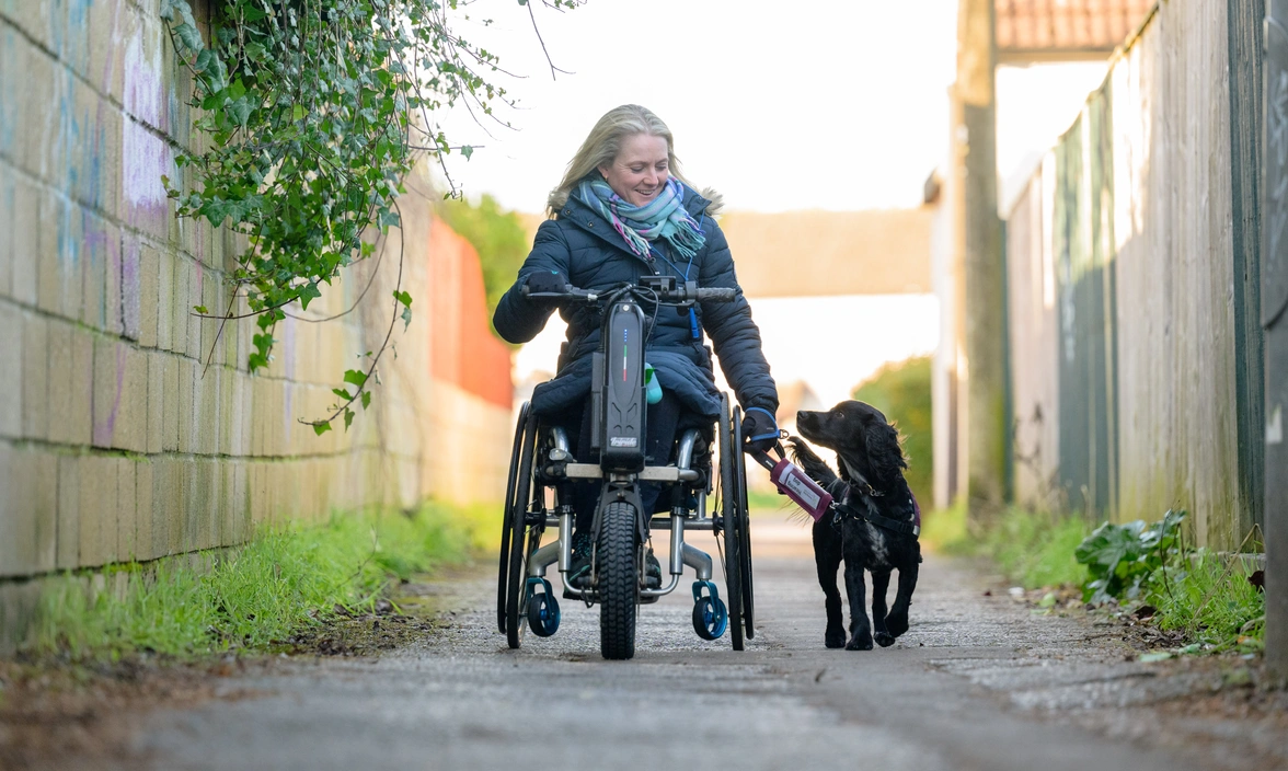 Woman in a wheelchair walking with a hearing dog on a lead down a path outside 