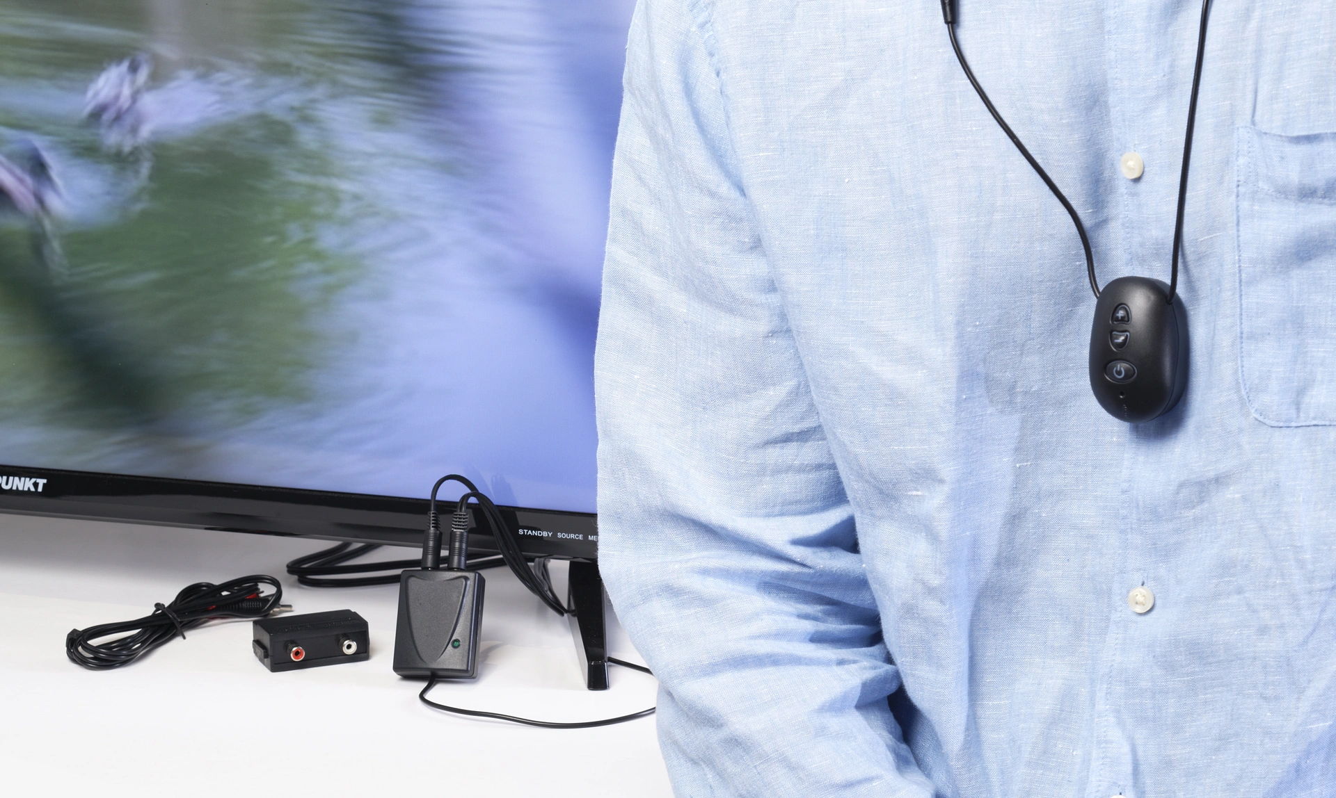 Close of of a person in a shirt wearing a hearing device around their neck with another device connected to the television behind them