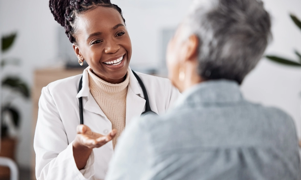 A medical professional in white coat with stethoscope smiling at another person and doing a hand gesture 