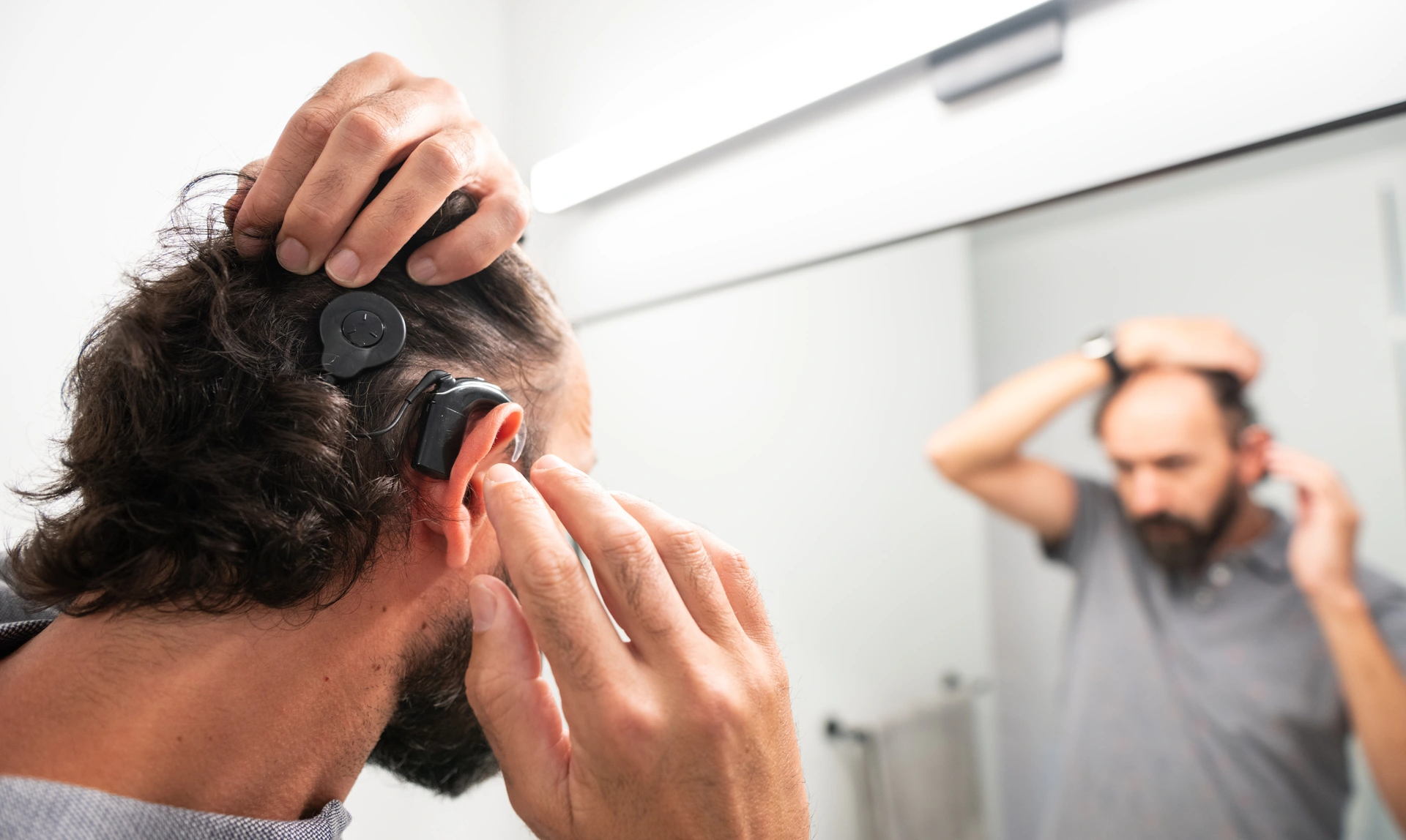 A man adjusting his cochlear implant in a room with a mirror so the back of his head and the reflection of his face is visible