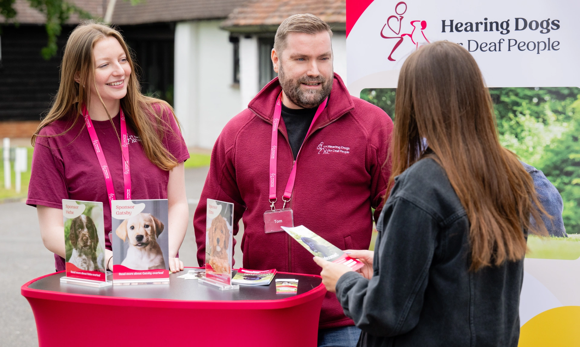 A man and 2 women are standing around a red table with brochures and pamphlets on it. Behind them is a banner that reads "Hearing Dogs for Deaf People" 