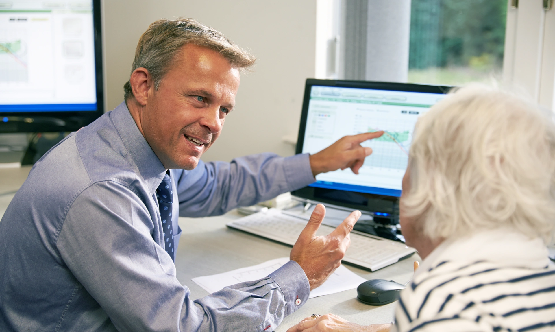 Audiologist sat looking at a patient while he smiles and points to a screen
