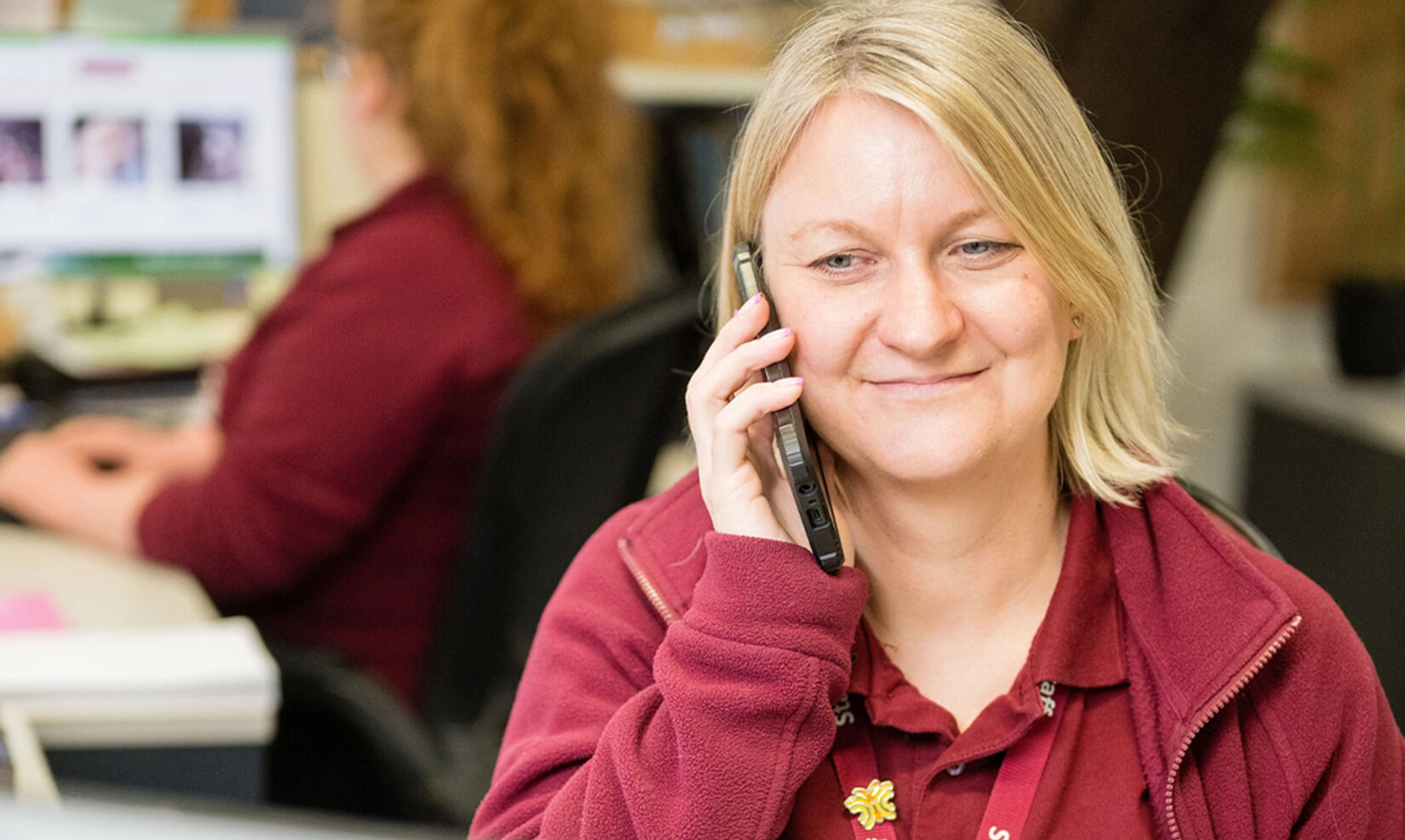 A woman in hearing dogs uniform sat at a desk smiling whilst on a mobile phone