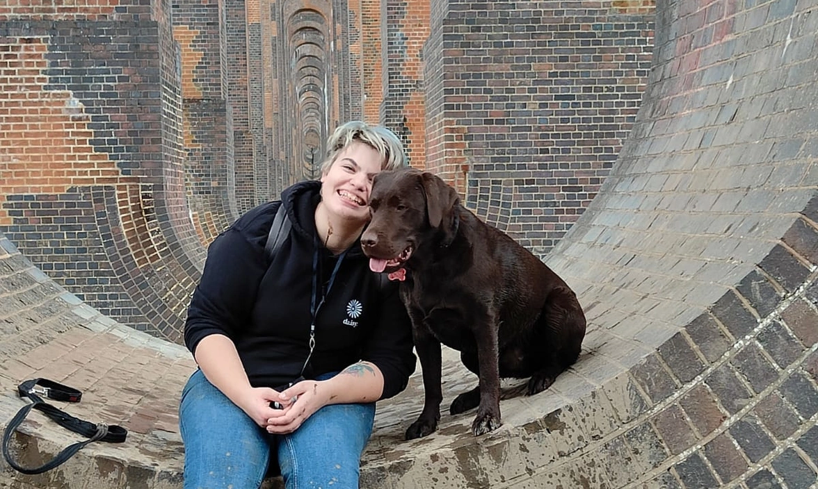 A smiling woman and a dark brown Labrador are sitting within a large brick edifice.